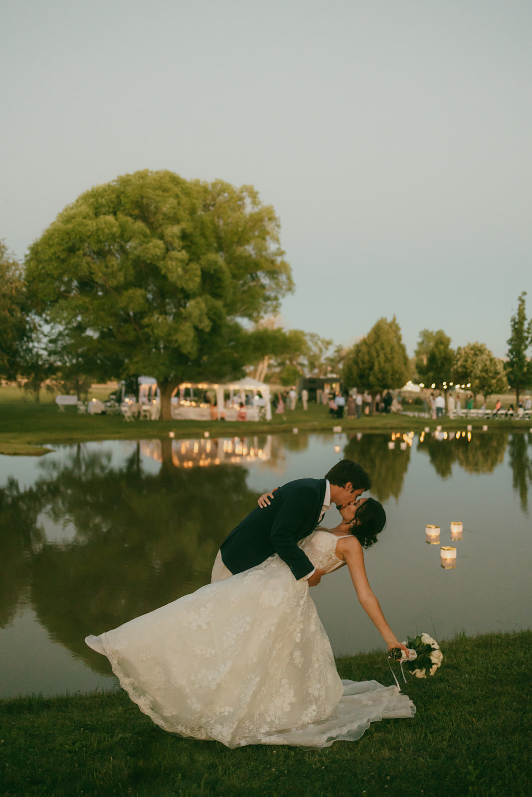 Married couple kissing in front of a pond at sunset with twinkly lights in the background