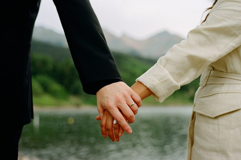 An up close picture of a couple's hands with their wedding rings in front of an alpine lake and mountains 