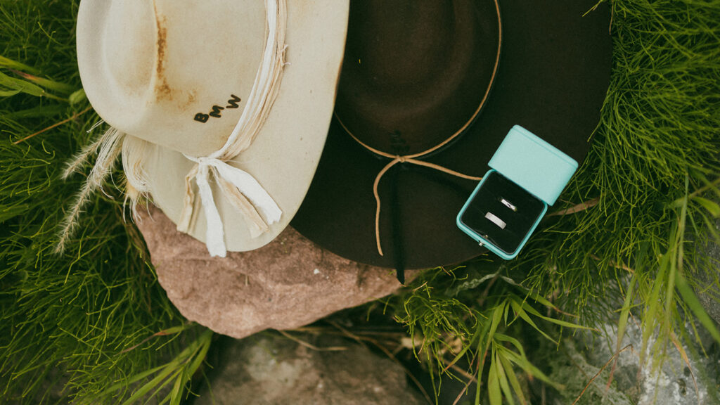 A pair of hats in the middle of a wildflower field with stamped initials.  A blue ring box with two wedding rings.