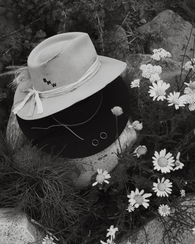 A pair of hats in the middle of a wildflower field with stamped initials and two wedding rings. 